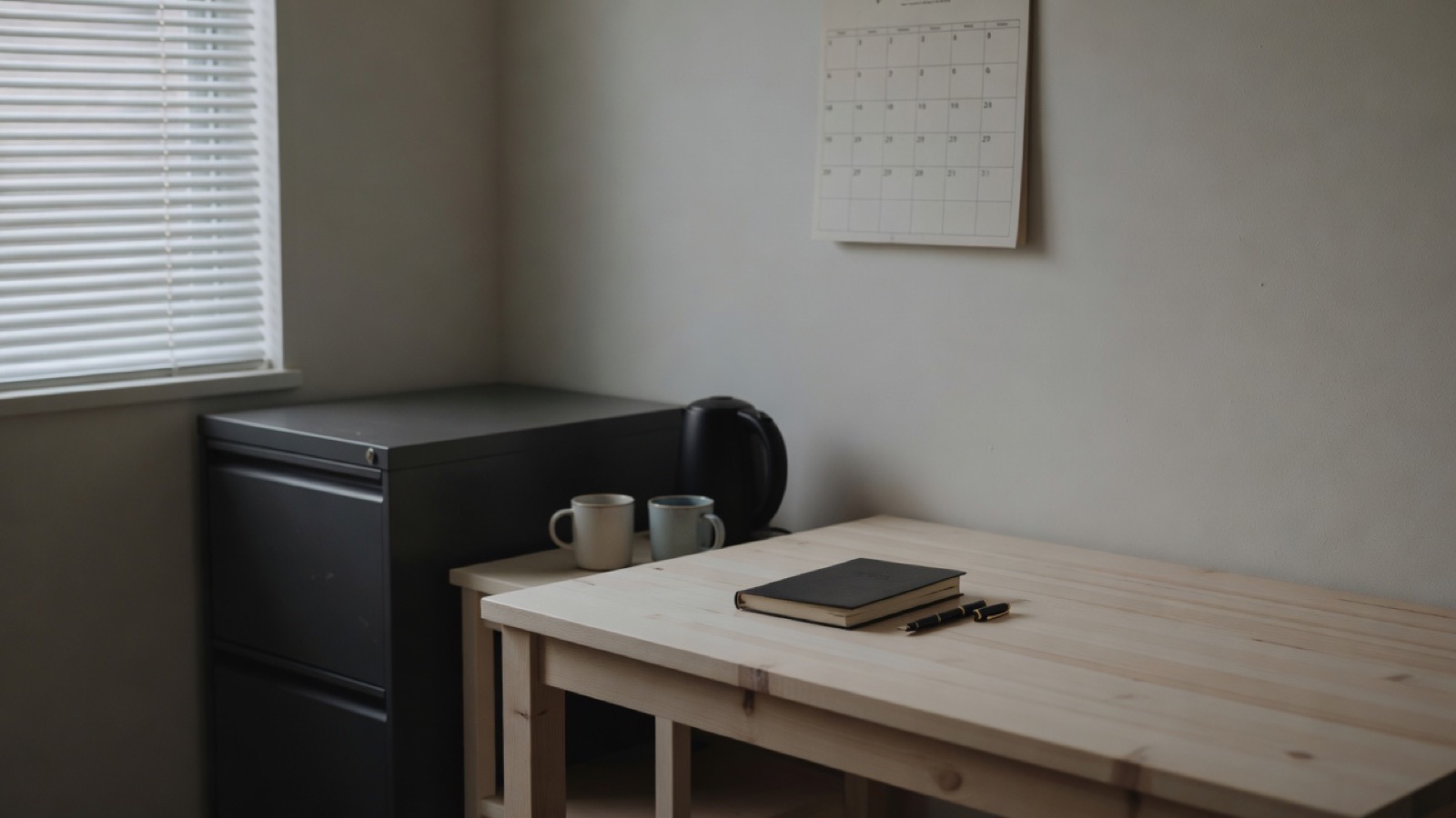 A small back office in the morning: a grey filing cabinet, a kettle on a side table, a wall calendar, a desk with a closed ledger and a mug, daylight through a blind